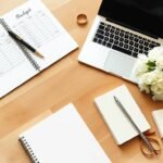 上から見た机のイメージ。ノートには英語で「Budget」と書かれており、鉛筆、花束、ノートパソコン、ハサミ、そして結婚指輪が置かれている。